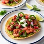 A crispy tortilla topped with guacamole, seasoned shrimp, diced tomatoes, onions, jalapeños, cilantro, and a dollop of sour cream, served with a lime wedge.