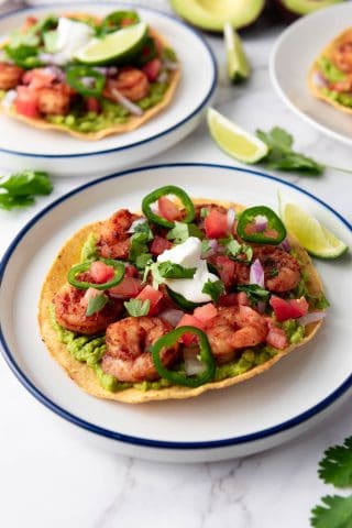 A crispy tortilla topped with guacamole, seasoned shrimp, diced tomatoes, onions, jalapeños, cilantro, and a dollop of sour cream, served with a lime wedge.