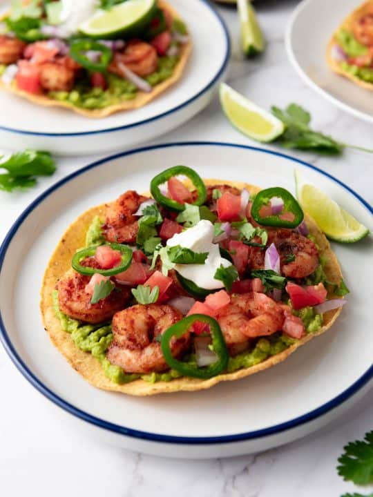 A crispy tortilla topped with guacamole, seasoned shrimp, diced tomatoes, onions, jalapeños, cilantro, and a dollop of sour cream, served with a lime wedge.