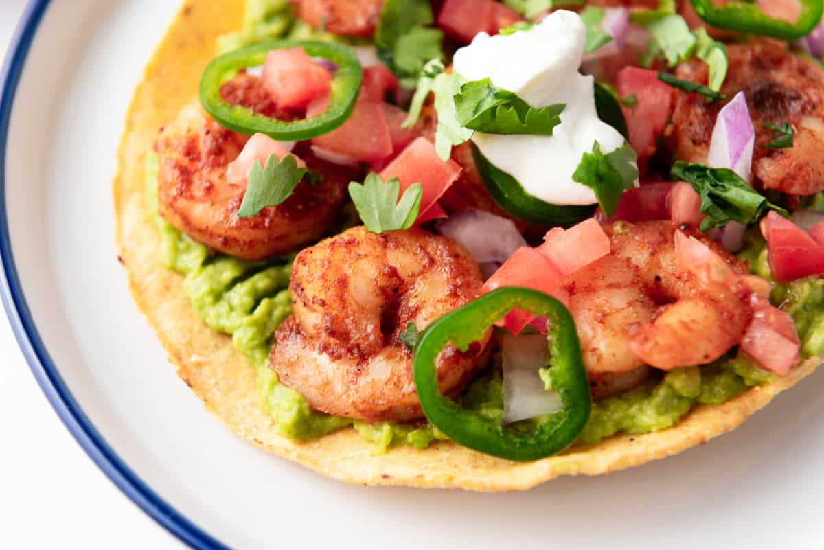 A tostada topped with guacamole, seasoned shrimp, sliced jalapeños, diced tomatoes, onions, cilantro, and a dollop of sour cream on a white plate.