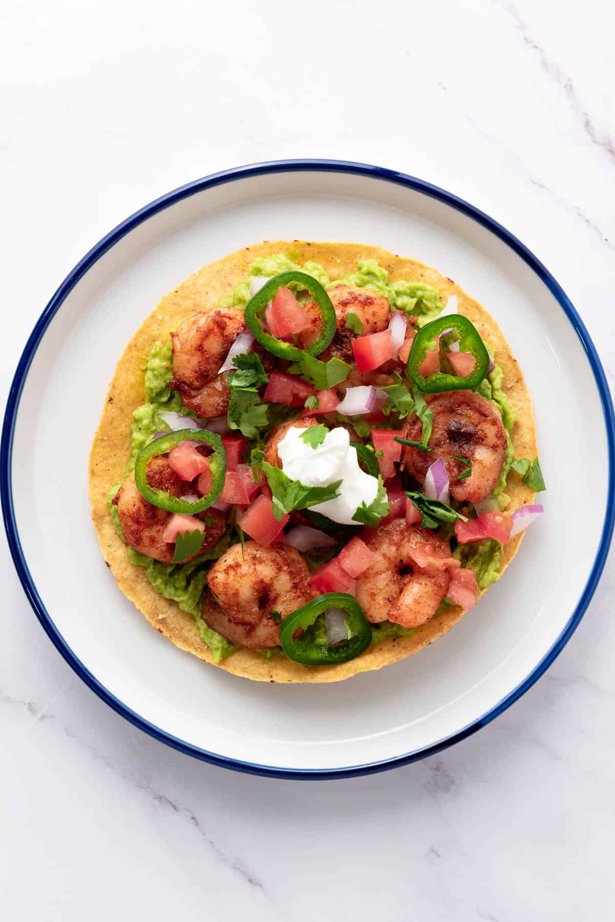 A tostada topped with shrimp, guacamole, sliced jalapeños, diced tomatoes, onions, cilantro, and a dollop of sour cream, served on a white plate with a blue rim.