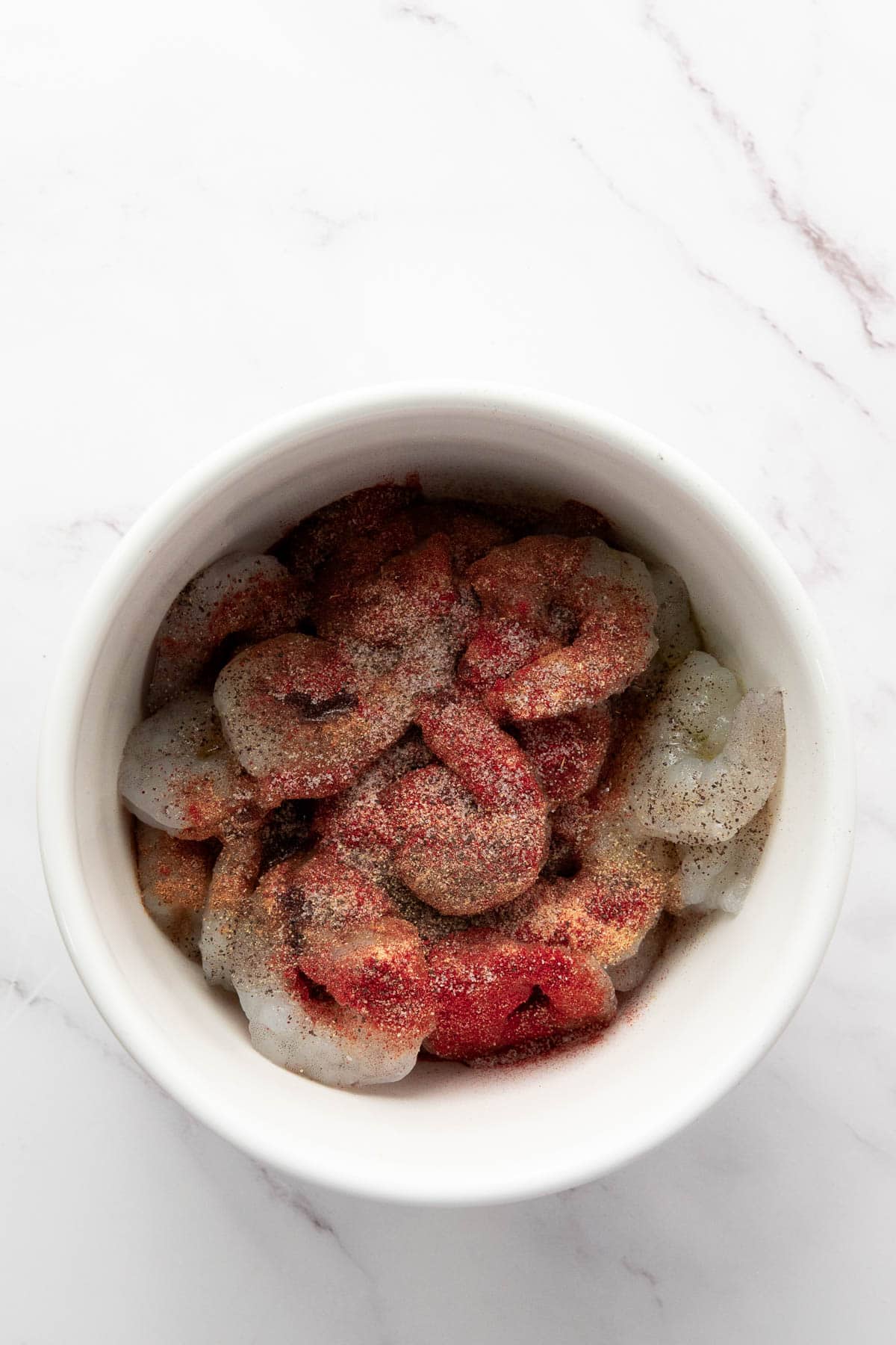 A white bowl filled with raw shrimp, seasoned with various ground spices, sits on a white marble surface.