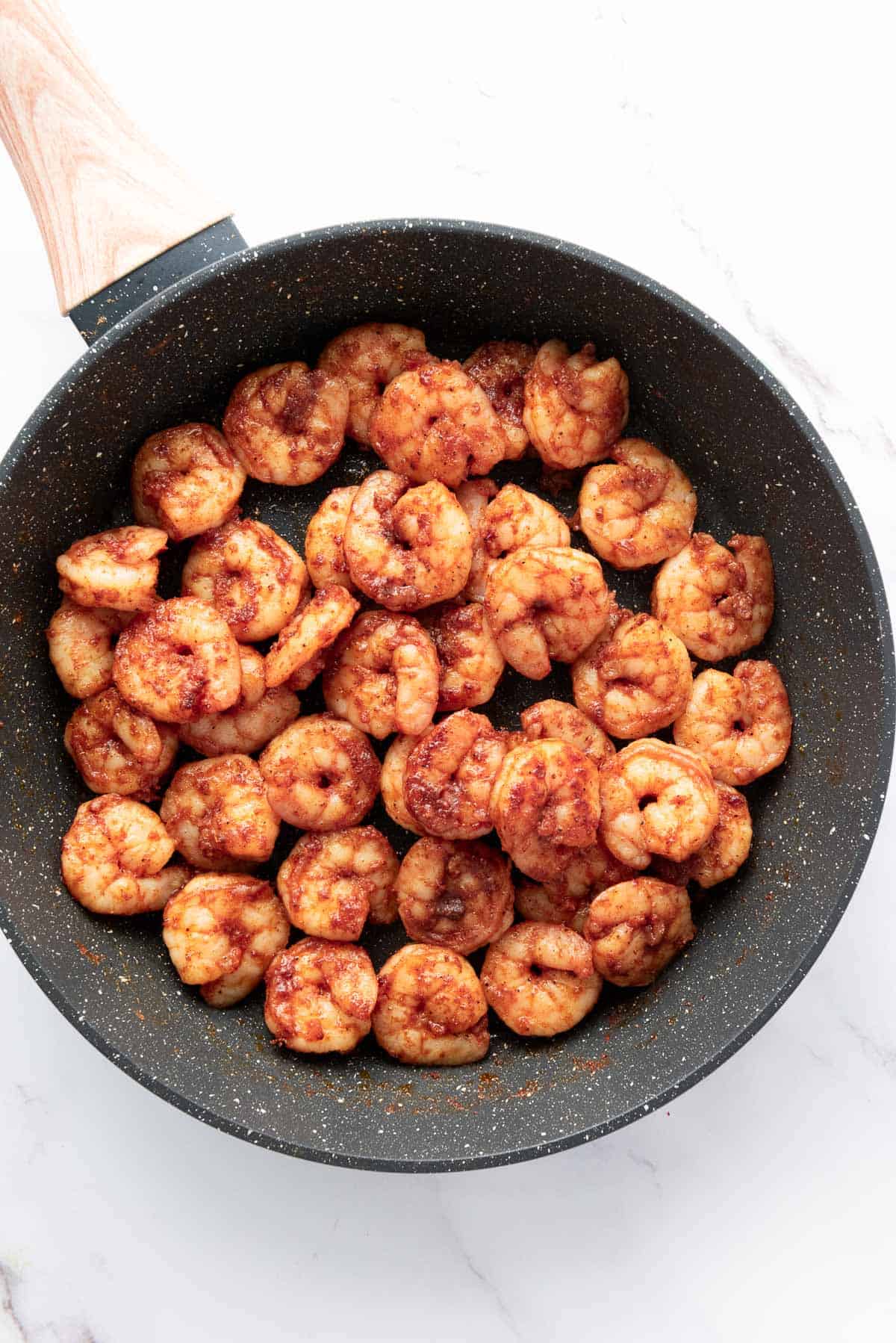 Cooked, seasoned shrimp in a black frying pan with a wooden handle, viewed from above on a white marble surface.