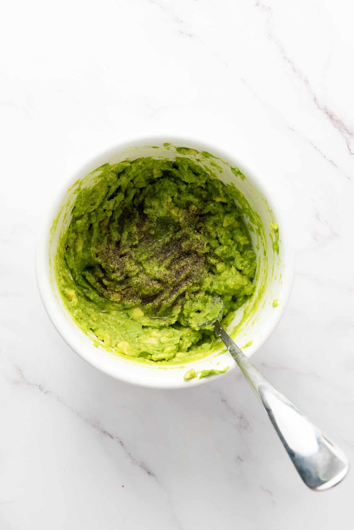 A white bowl of mashed avocado with black pepper on top, and a metal spoon resting inside, on a white marble surface.