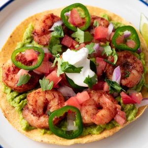 A tostada topped with guacamole, grilled shrimp, sliced jalapeños, diced tomatoes, onions, cilantro, and a dollop of sour cream is served on a white plate with a lime wedge.